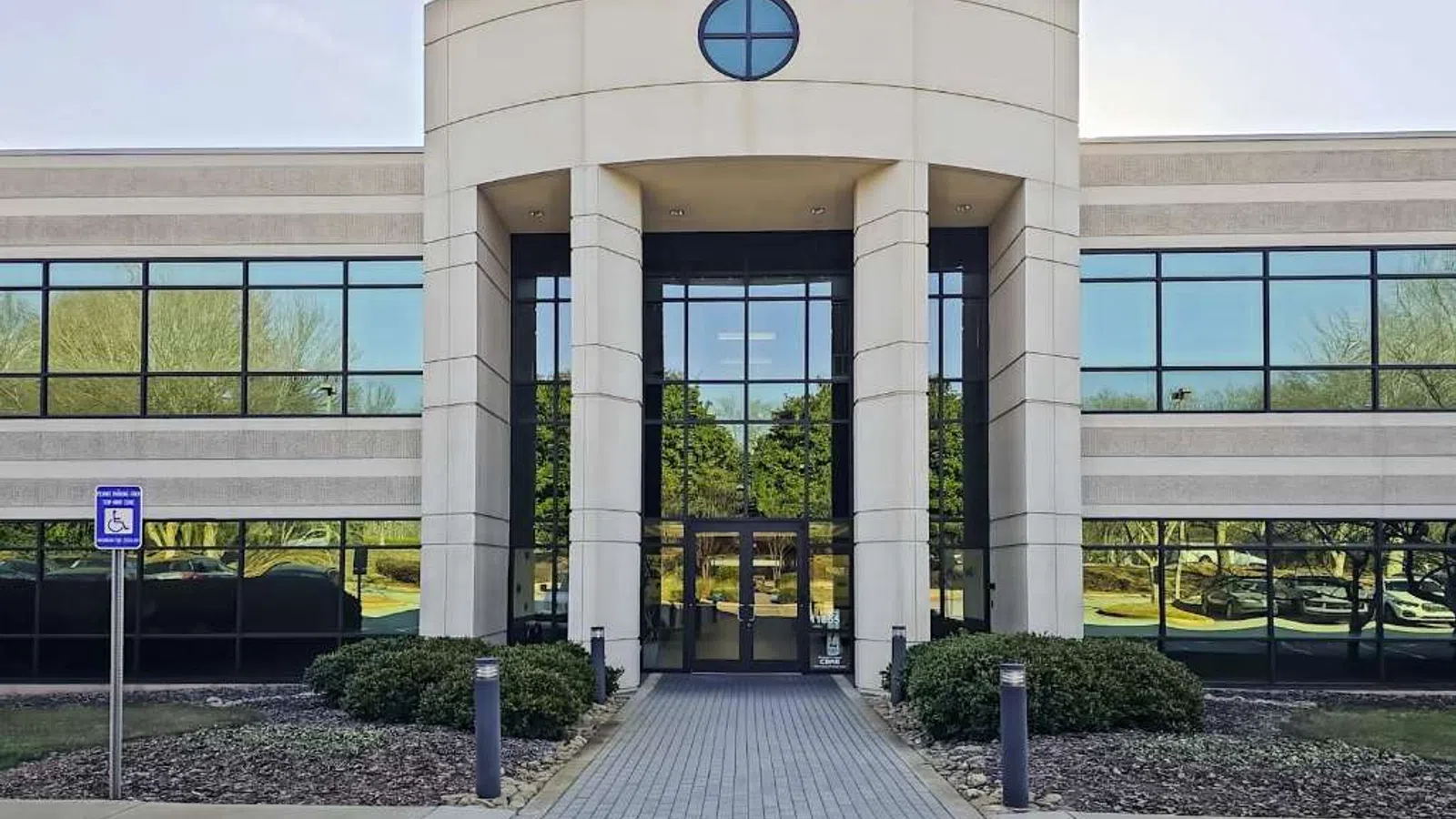Modern office building entrance with large glass windows, stone pillars, and landscaped greenery in front.