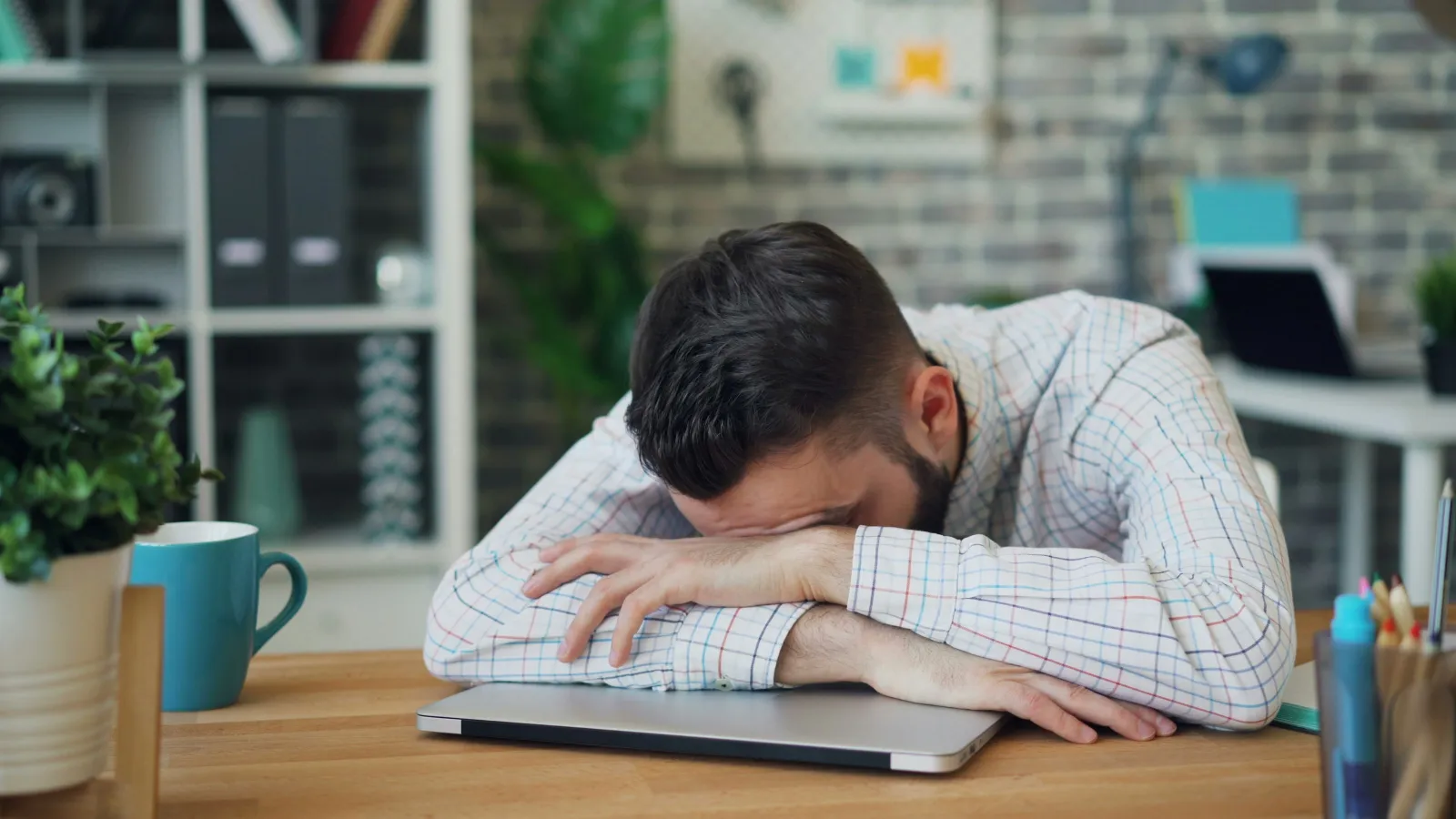 Man resting his head on folded arms over closed laptop on a desk looking stressed in a home office.