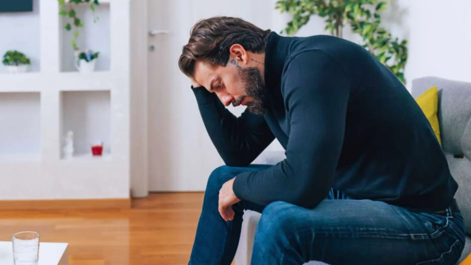 Man sitting on a sofa with his head in his hand, appearing stressed or deep in thought in a living room.