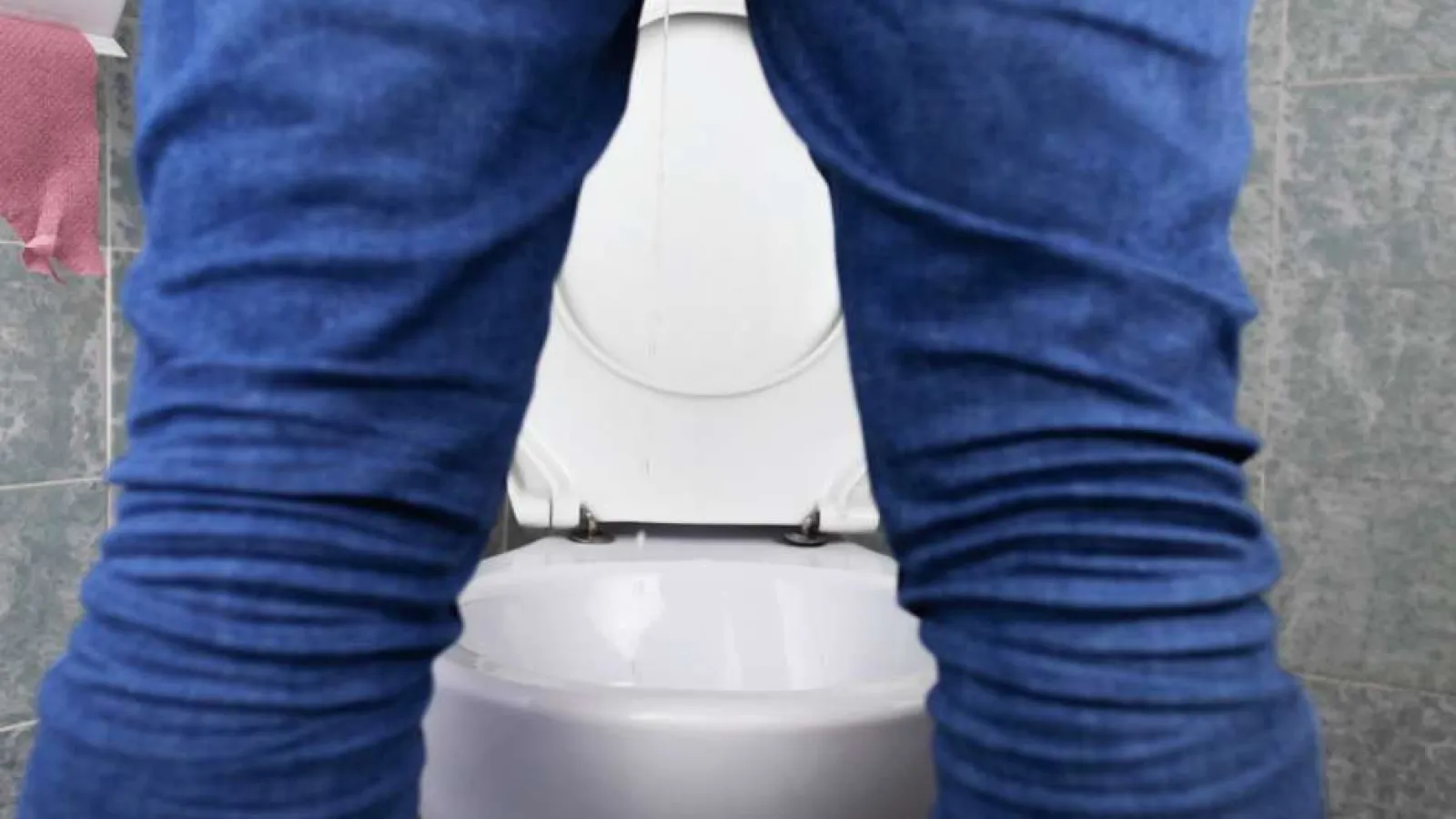 Person standing in front of a white toilet in a tiled bathroom, wearing blue jeans, viewed from behind.