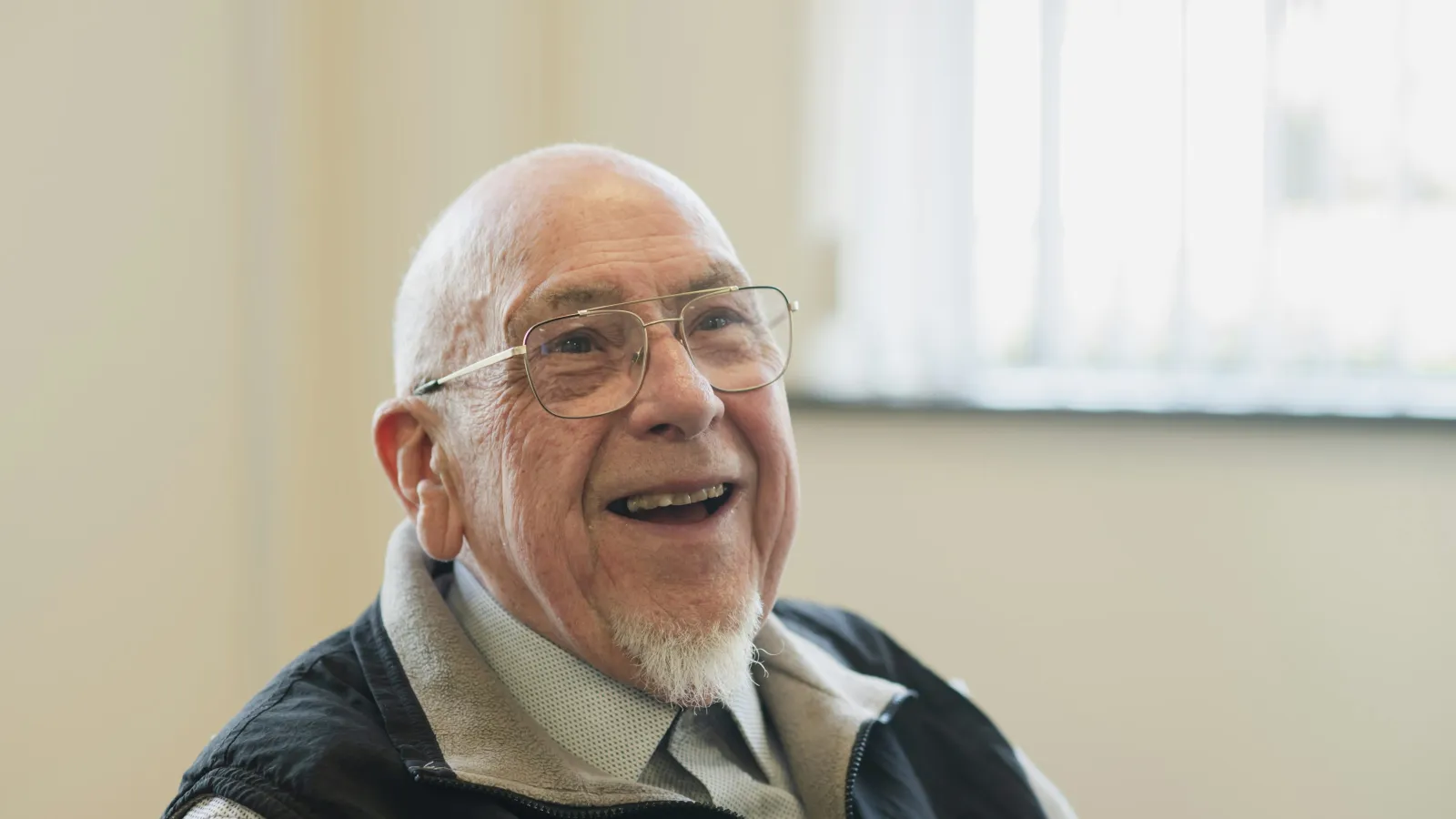 Happy elderly man with glasses and a beard smiling indoors by a bright window, wearing a black vest and shirt.