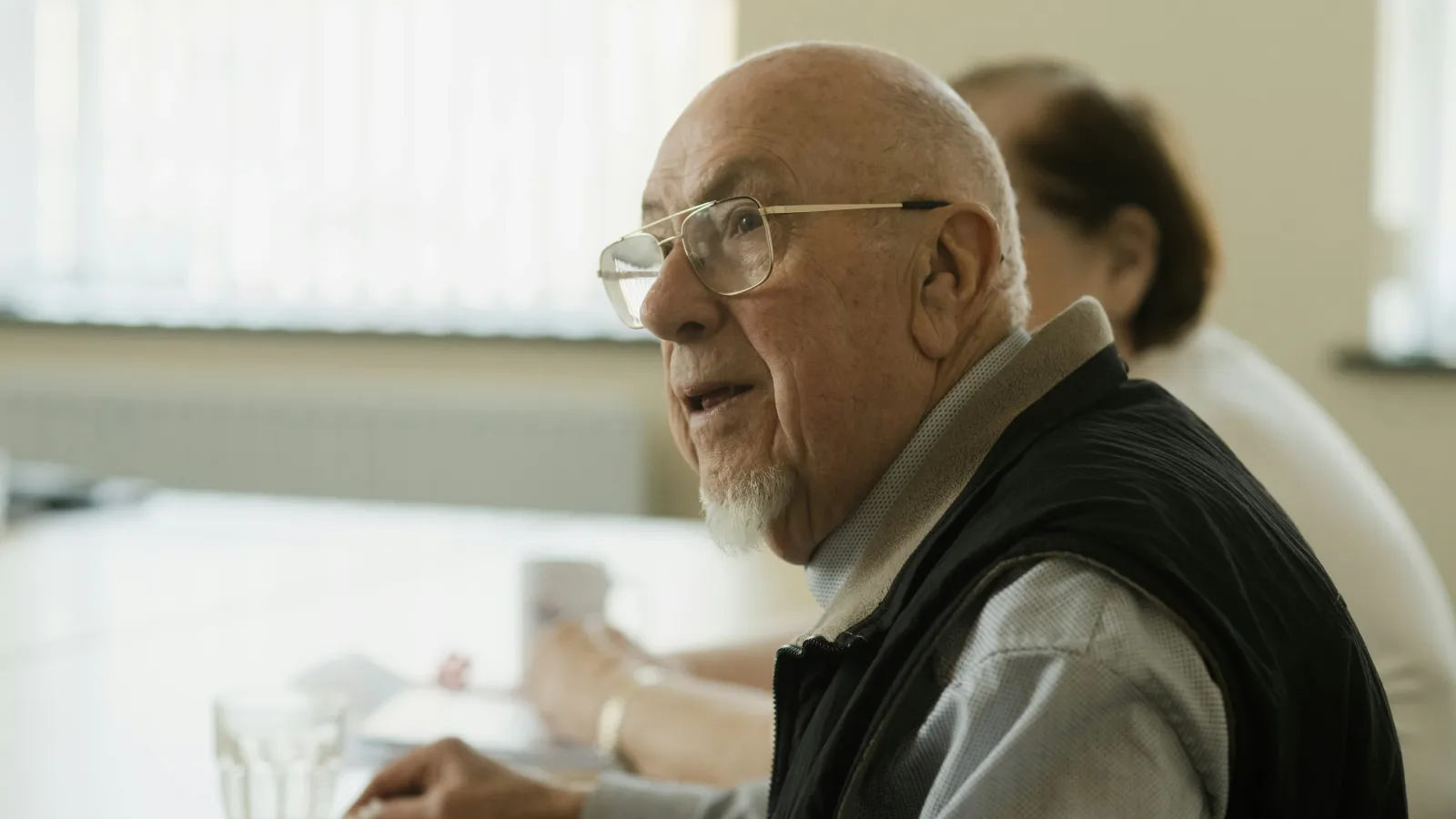 Elderly man with glasses sitting at a table, holding a fork with food, engaging in conversation indoors.