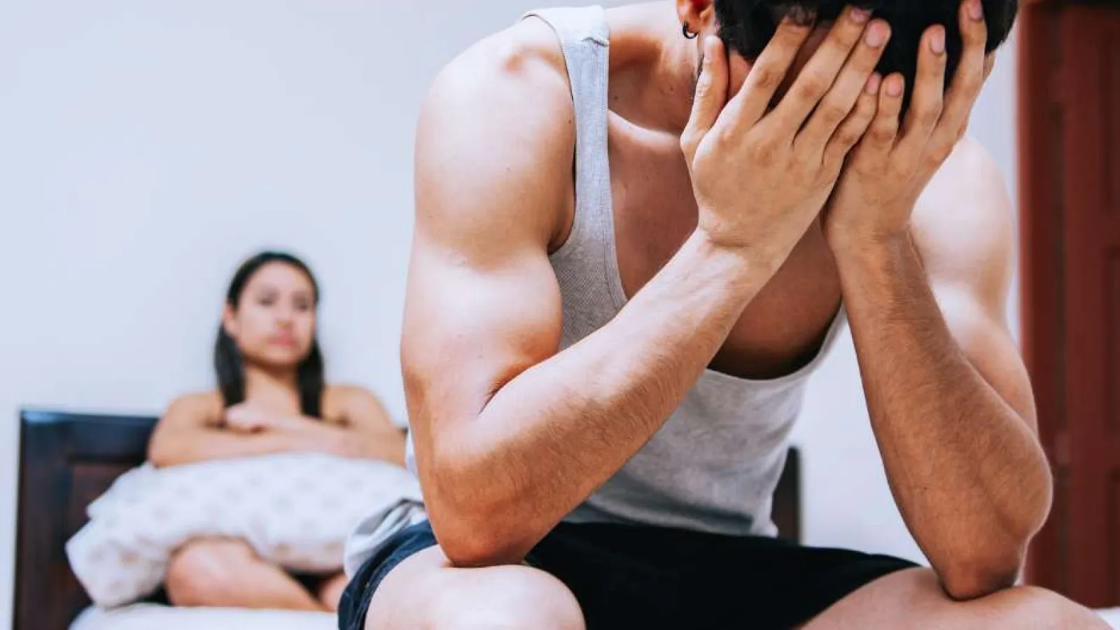Man sitting on bed covering his face in distress while woman sits in background holding pillow.