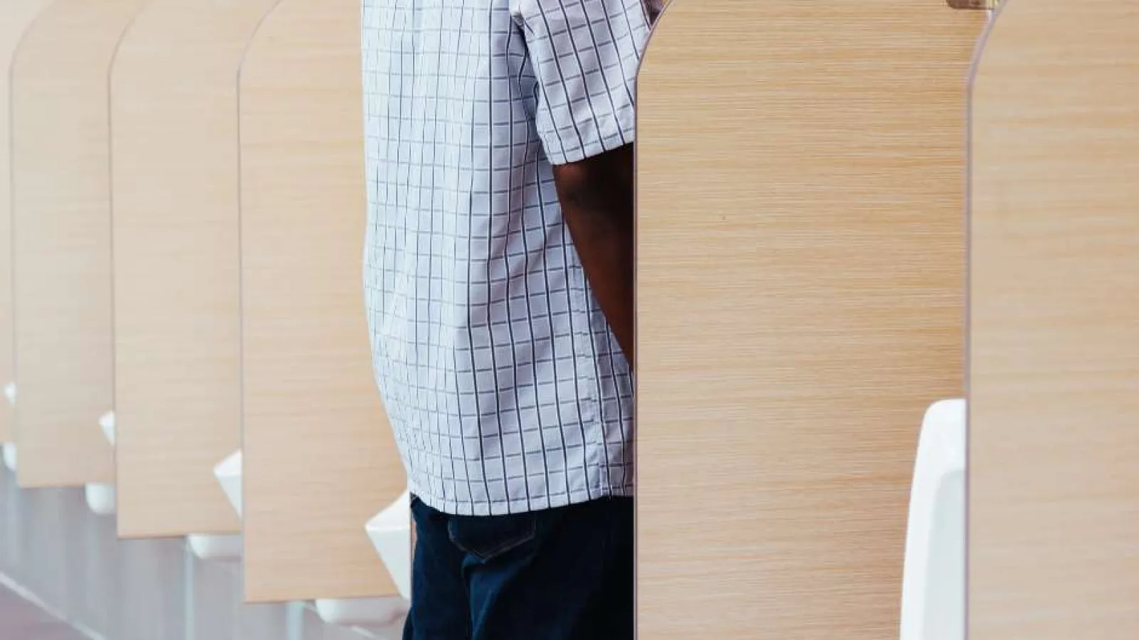 Man wearing a checkered shirt using a urinal in a restroom with wooden privacy dividers.