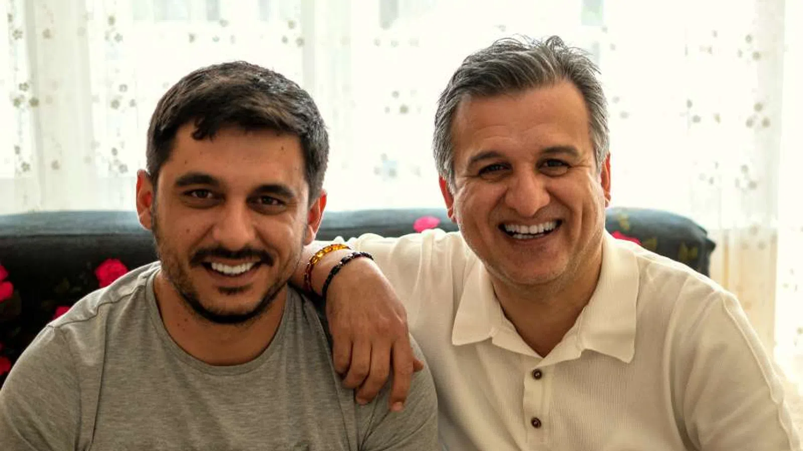 Two men smiling together indoors, one with arm around the other's shoulder, sitting on a couch by a window.