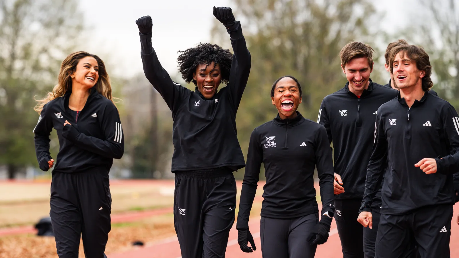 Five diverse athletes in black Adidas tracksuits smiling and celebrating on a running track outdoors.