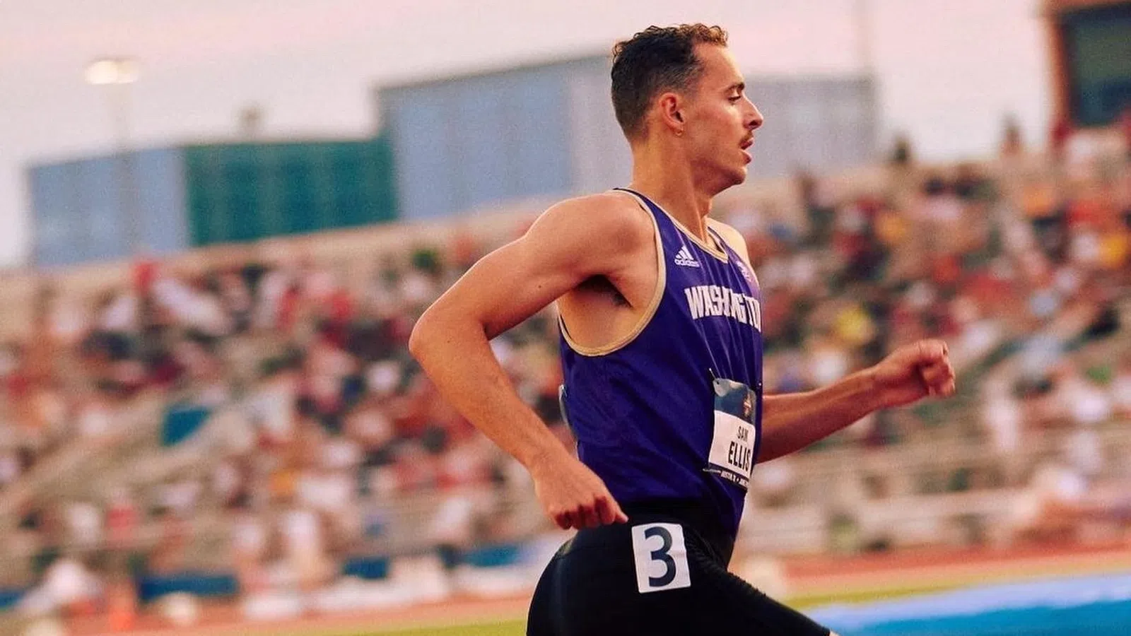 Male athlete in purple Washington jersey sprinting on a red track during a race with blurred spectators in background.