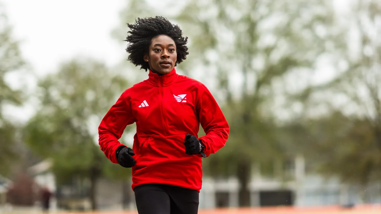 Athlete running on an outdoor track wearing a red jacket and black leggings during daytime training.