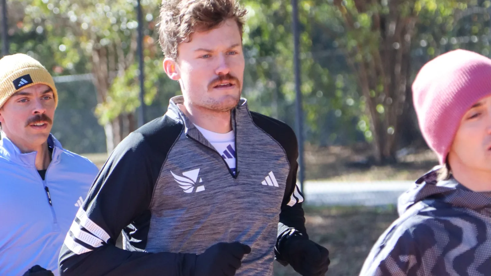 Three men jogging outdoors in cold weather wearing gloves, athletic wear, and hats in a park with trees.