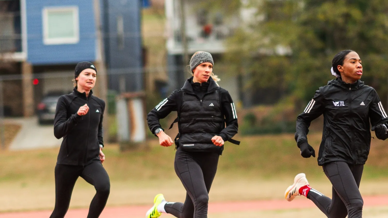 Four women running on an outdoor track in athletic clothing during daytime with houses and trees in the background.