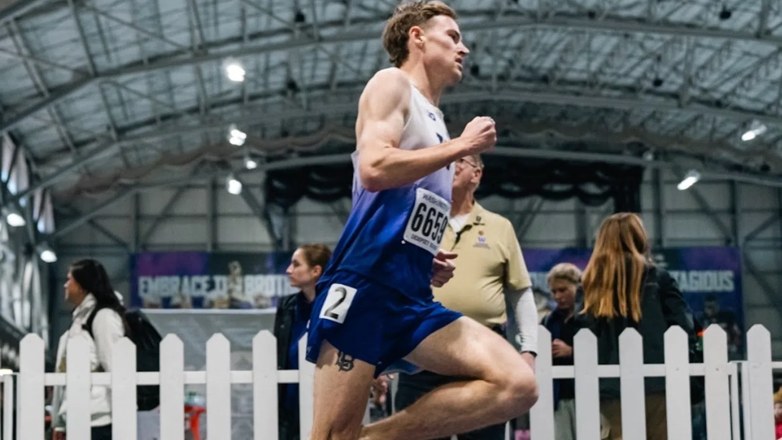 Male athlete in blue running uniform sprinting on indoor track with spectators behind a white fence.