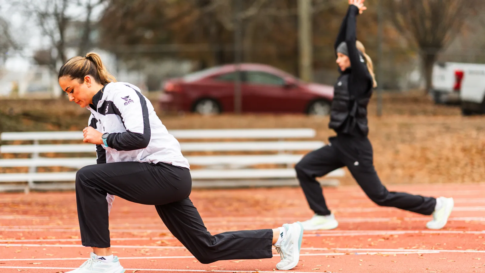 Two women performing lunges on an outdoor running track during autumn training session.