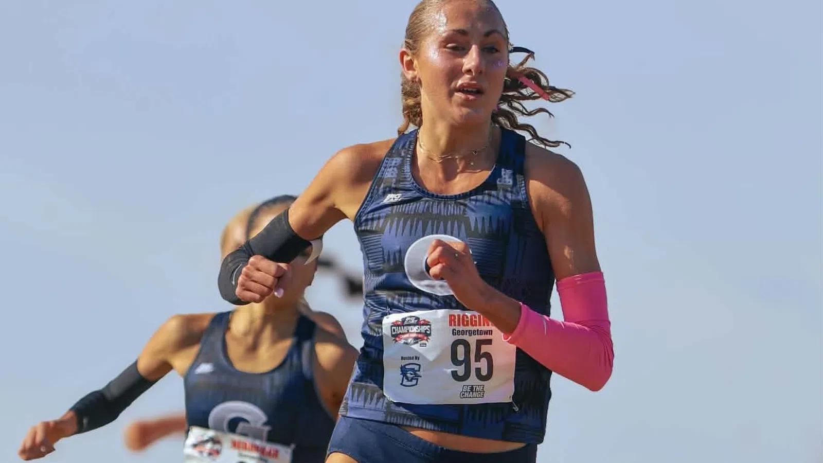 Two female cross country runners racing outdoors on a grassy trail in athletic gear under clear sky.
