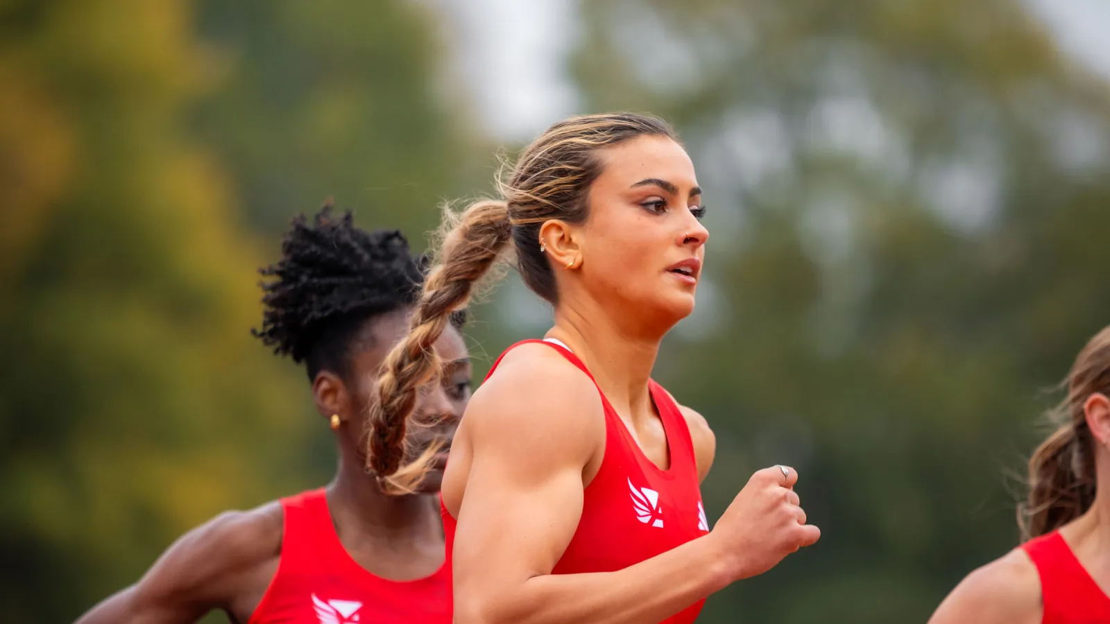 Female athletes wearing red sportswear sprinting outdoors on a track with a blurred natural background.