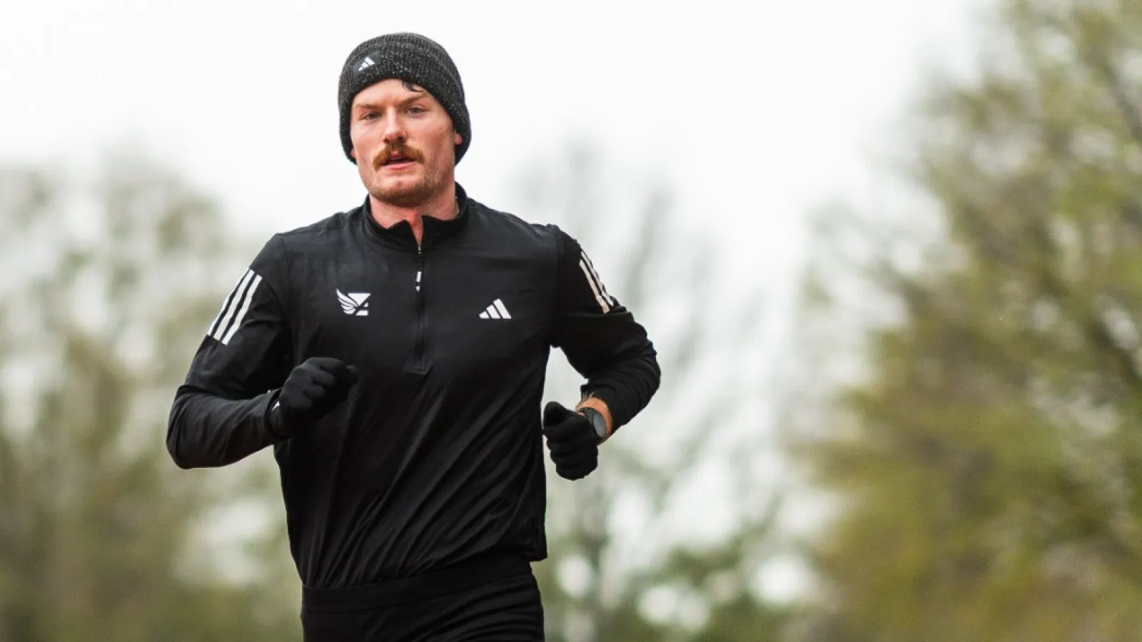 Male athlete in black running gear sprinting on an outdoor red track with trees in background
