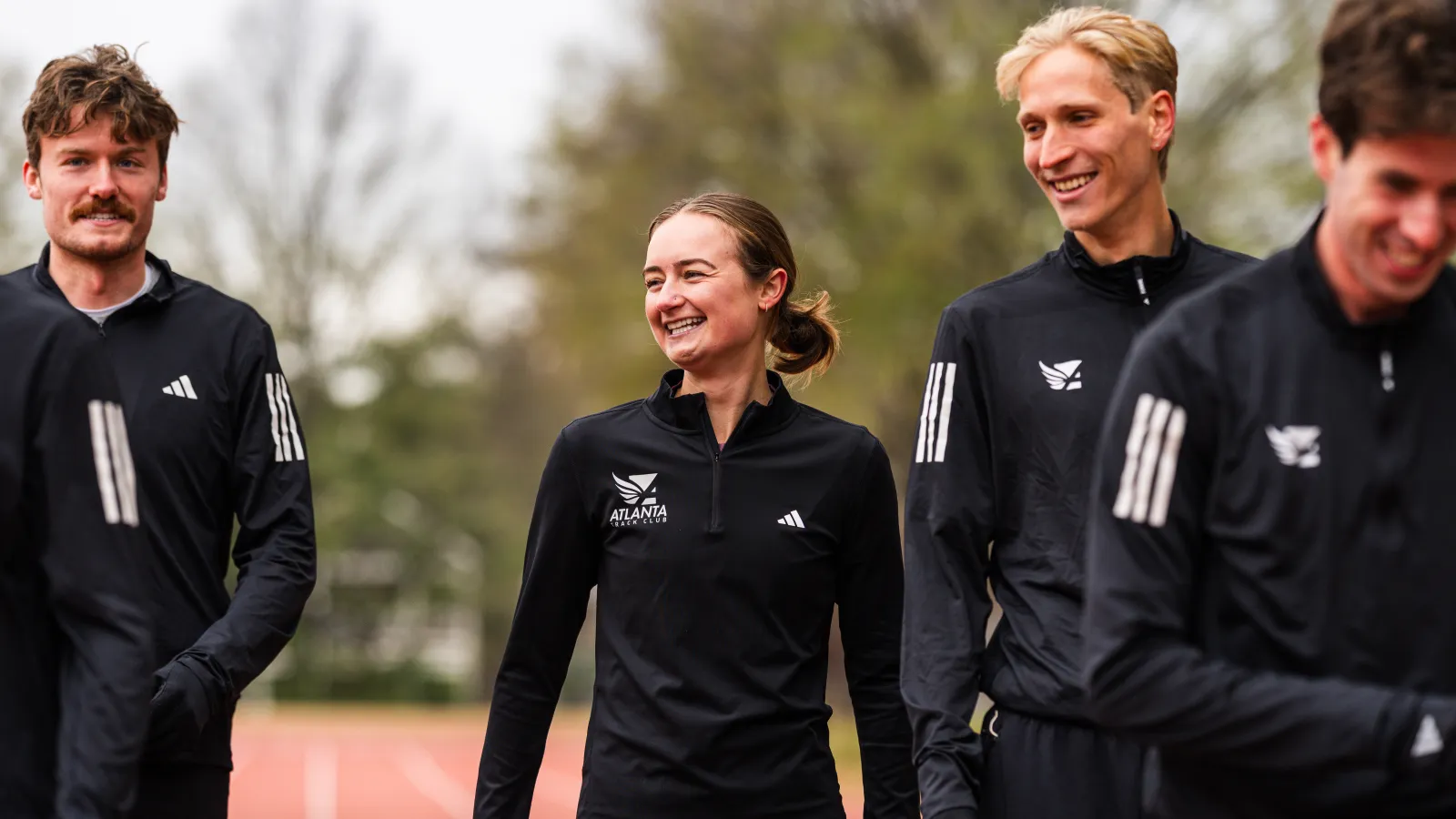 Four young athletes wearing black Atlanta Track Club jackets smiling and standing on a running track outdoors.