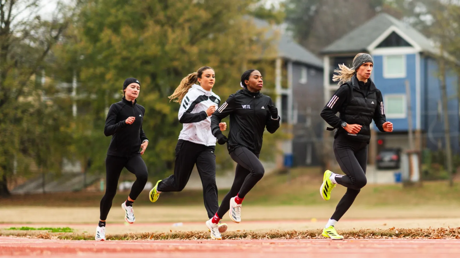 Four women running on an outdoor track in cold weather with houses and autumn trees in the background