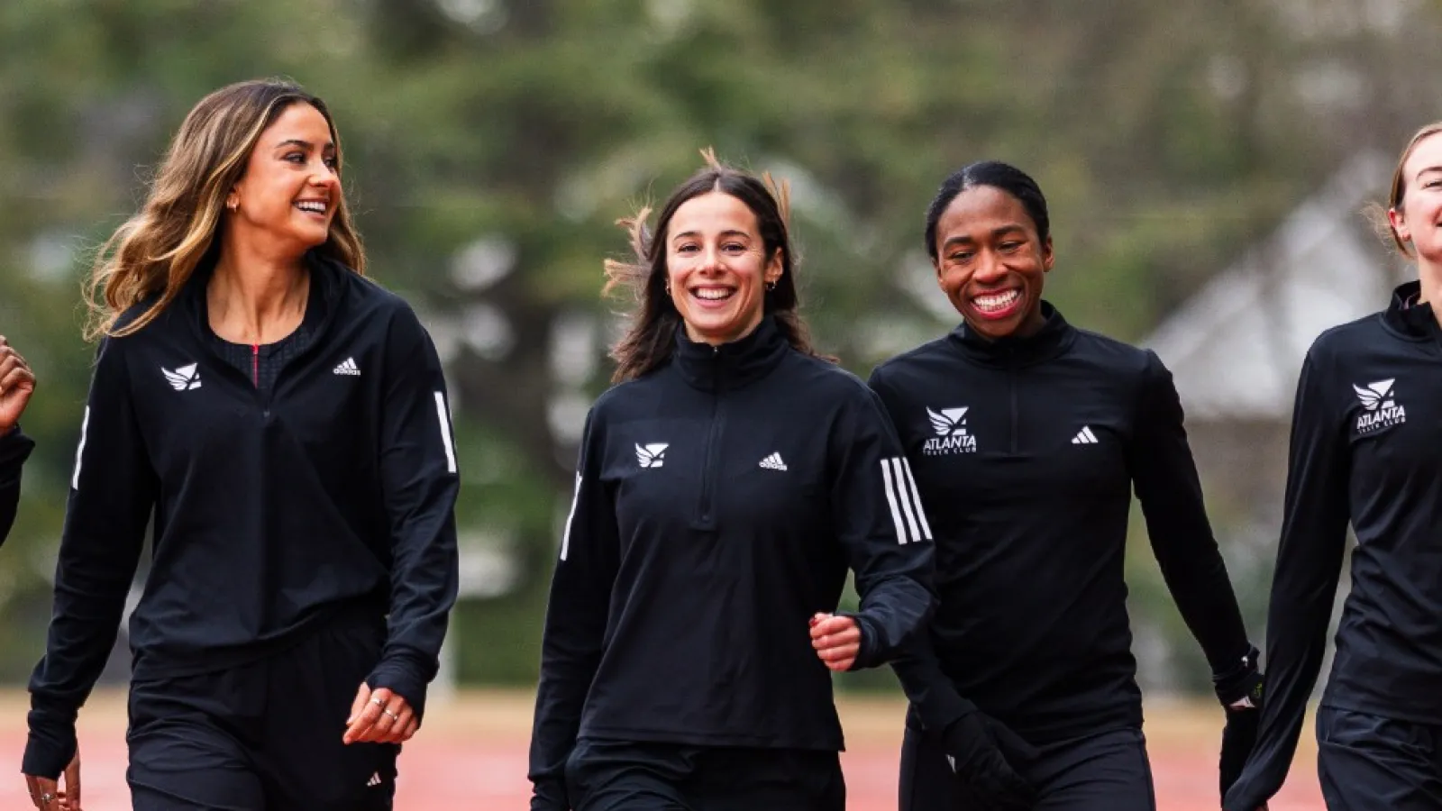 Six women athletes walking together on an outdoor track wearing black sportswear, smiling and enjoying each other's company.