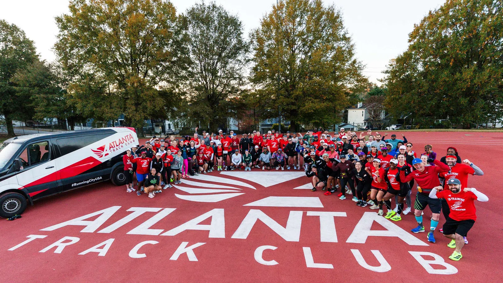 Large group of people in red shirts posing on Atlanta Track Club logo on running track with branded van nearby