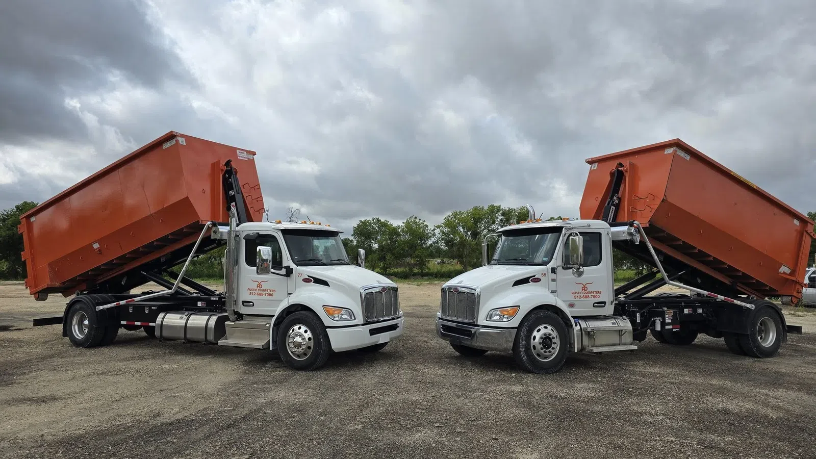 Two white dump trucks with raised orange containers parked on a gravel lot under a cloudy sky.