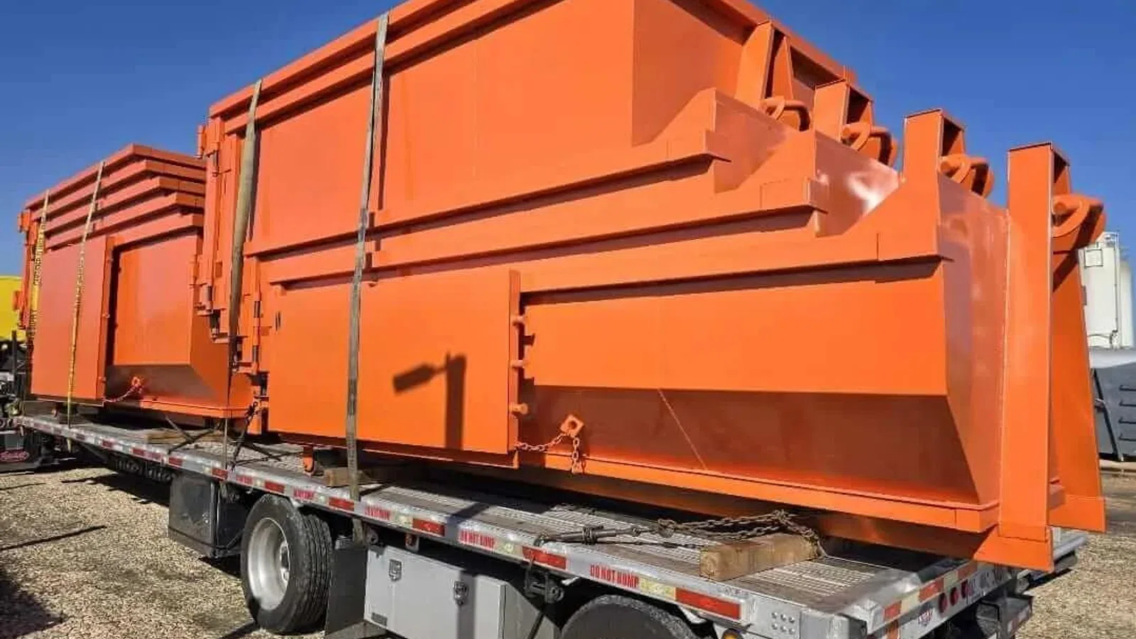 Bright orange industrial scrap metal containers secured on a flatbed trailer under clear blue sky.