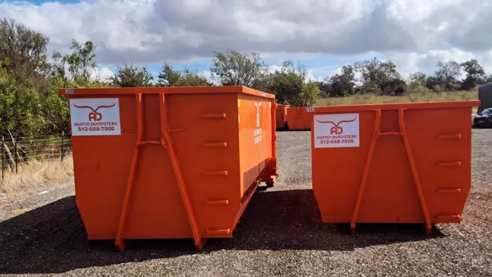 Two large orange dumpsters on gravel lot under blue sky with clouds and trees in background.