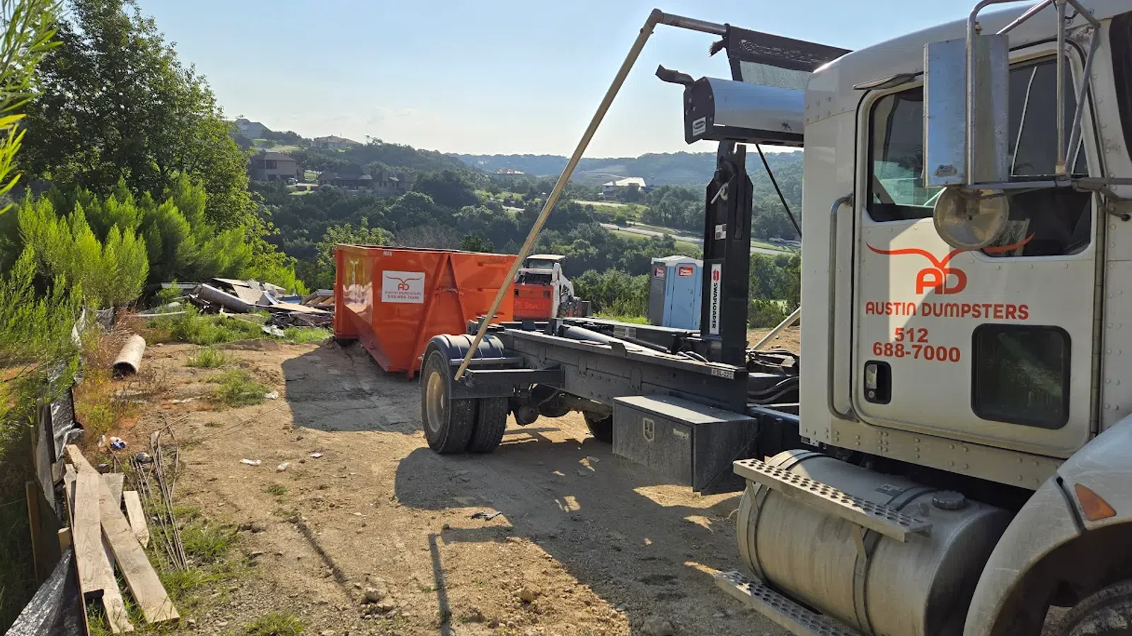 Austin Dumpsters truck unloading orange dumpster at a rural site with hills and greenery in the background.