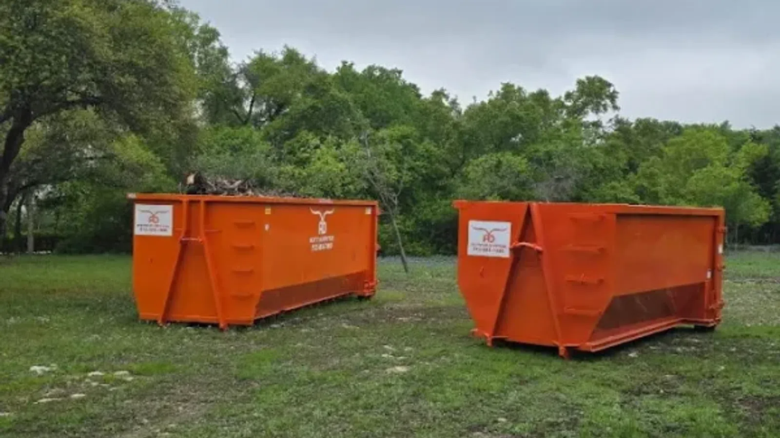 Two large orange construction dumpsters on grassy area with trees and cloudy sky in background