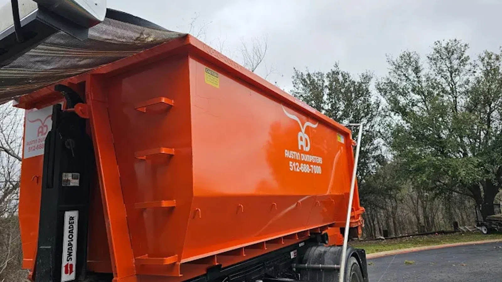 Large orange dumpster trailer with Austin Dumpsters branding parked on a driveway near trees and cloudy sky.