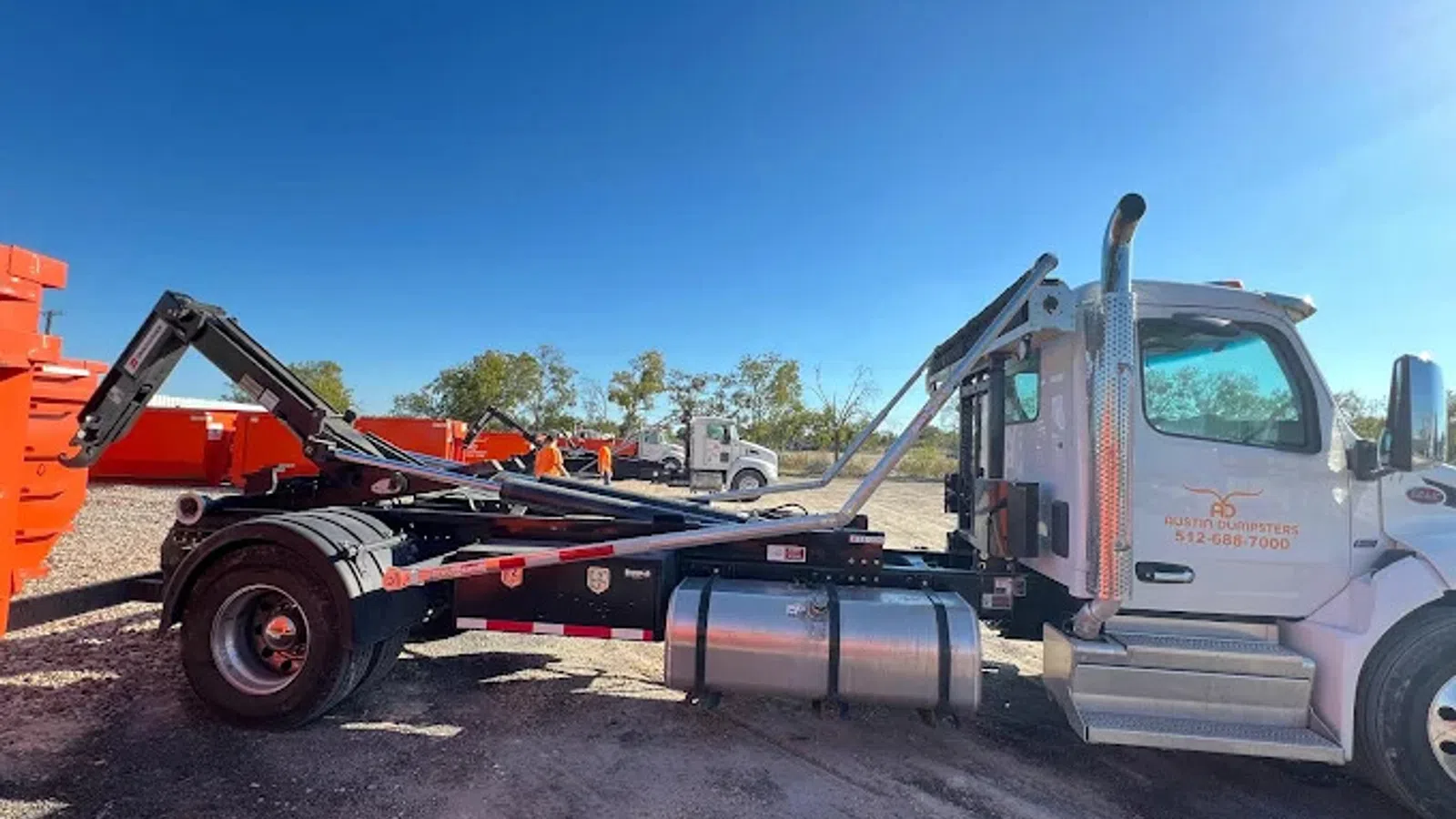 White roll-off truck with hydraulic arm parked near orange containers under clear blue sky.
