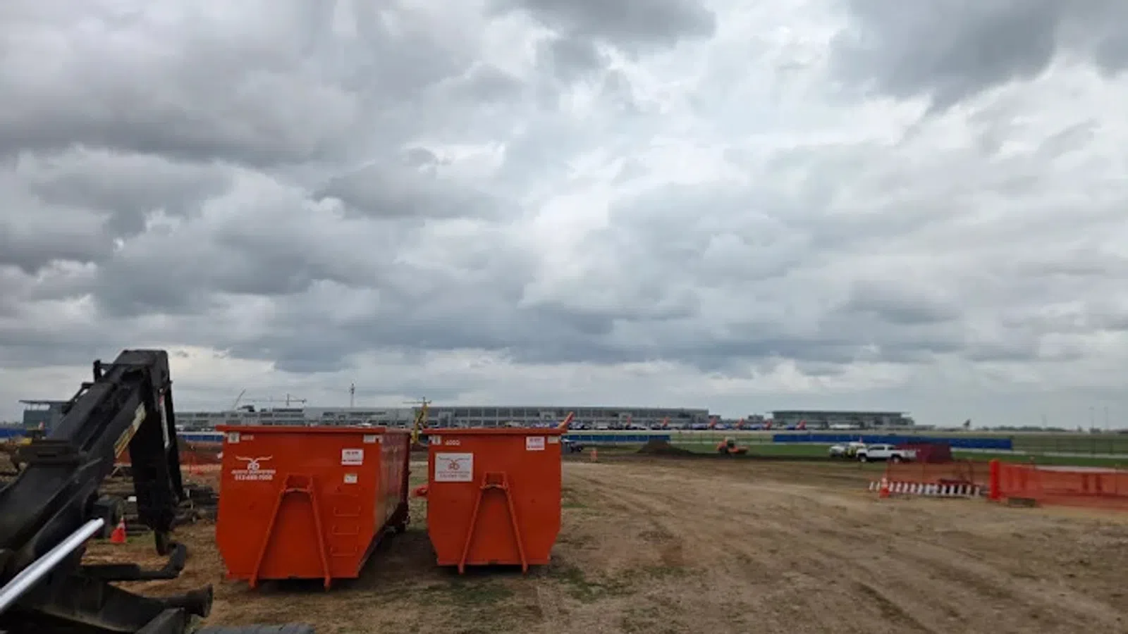 Construction site with two orange dumpsters under cloudy sky on a dirt field near industrial buildings.