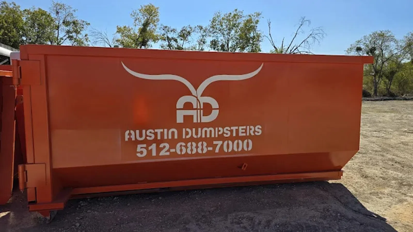 Large orange dumpster labeled Austin Dumpsters with phone number 512-688-7000 outdoors on dirt with trees