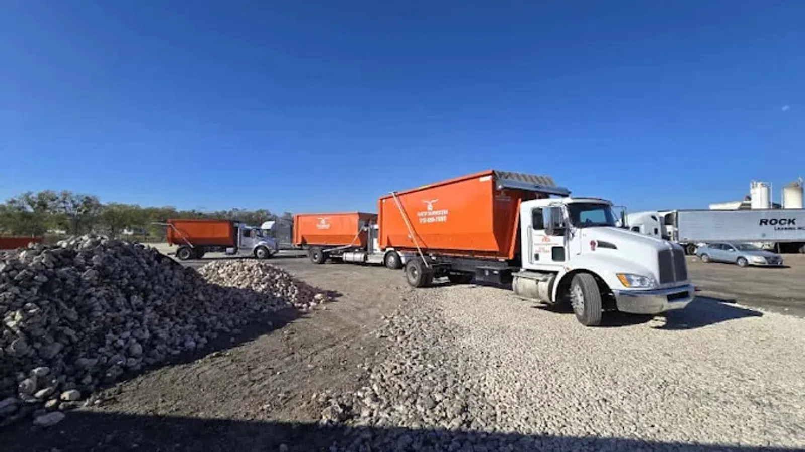 White and orange dump trucks parked on gravel near large piles of rocks under a clear blue sky.