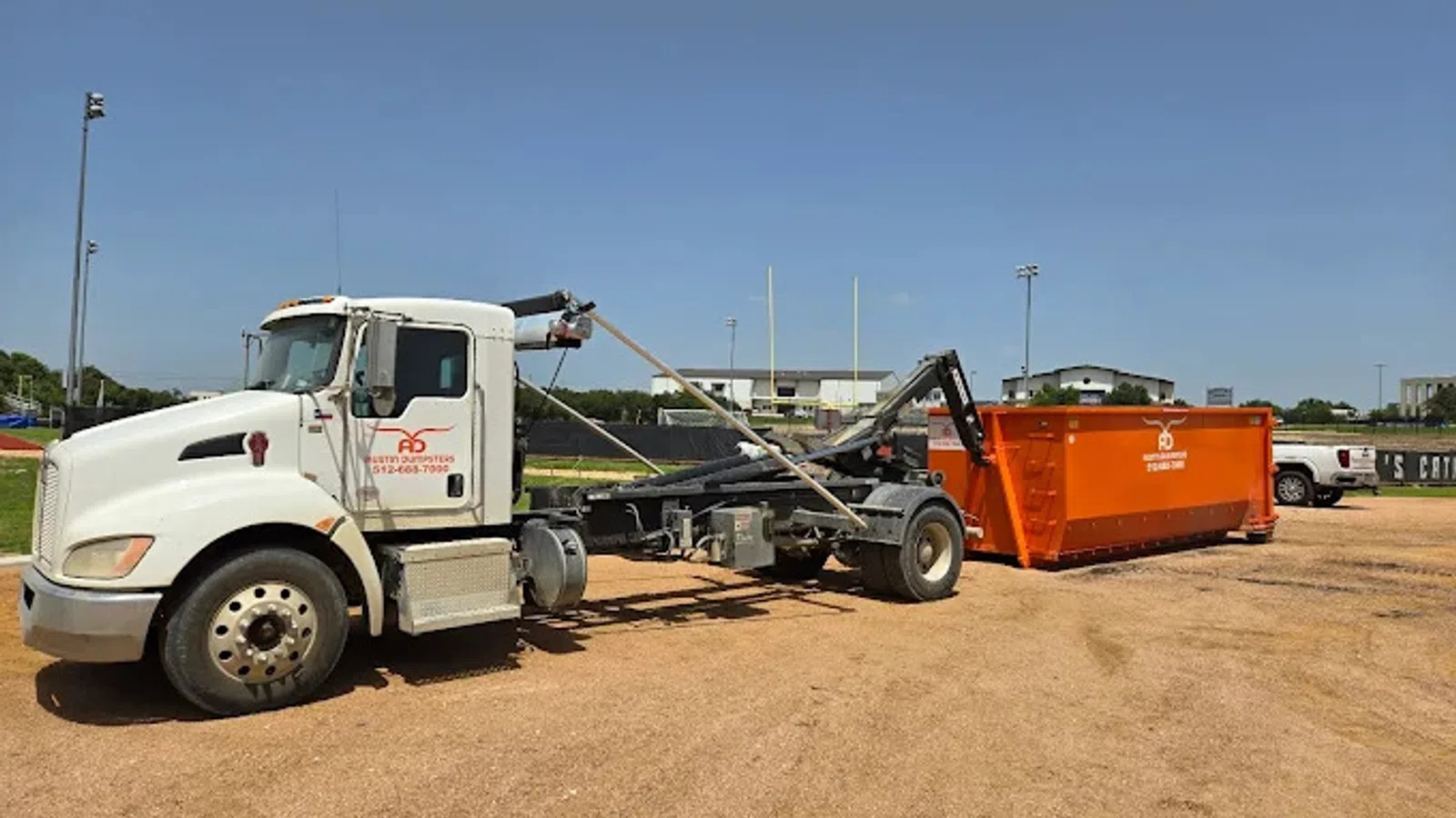 White roll-off truck attached to an orange dumpster on a dirt lot under clear blue sky