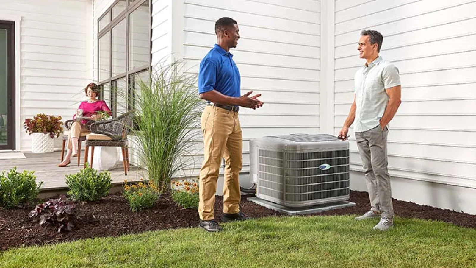 Two men discussing an air conditioning unit in a well-maintained yard, with a woman relaxing nearby.