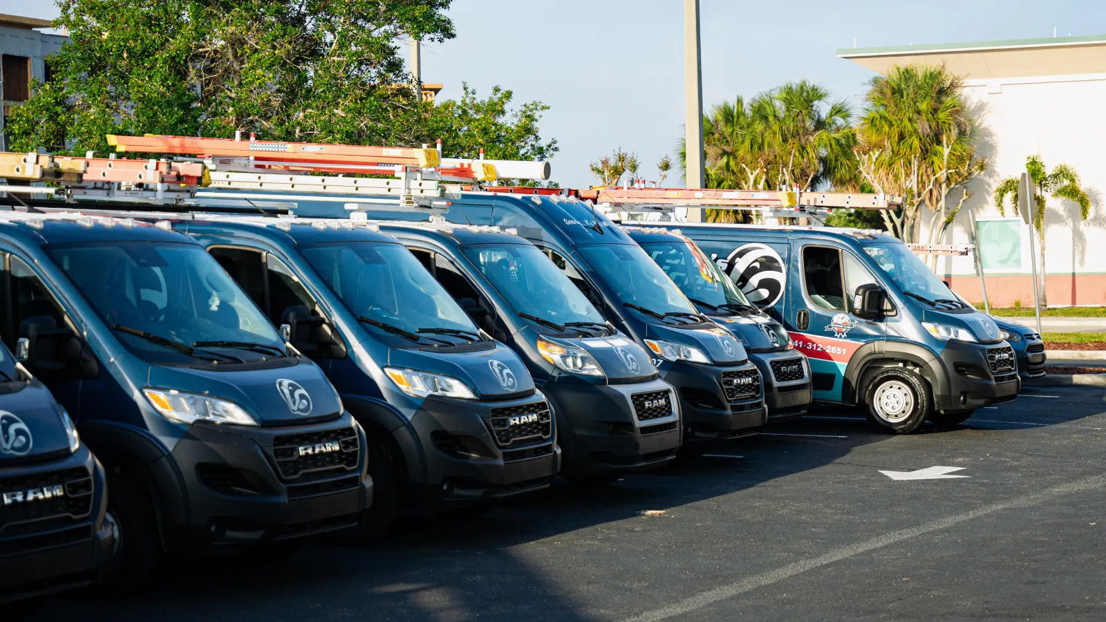 Row of black RAM service vans with ladders parked outdoors in a sunny lot with trees and buildings in the background