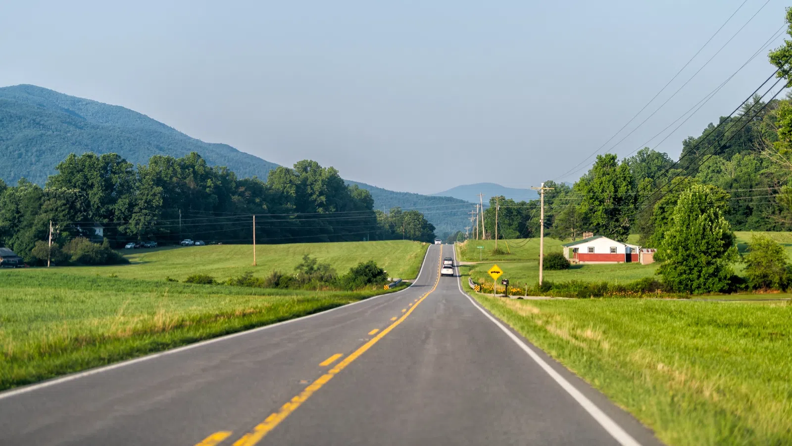 Two-lane rural road stretches toward mountains with green fields and a house on the side under a clear sky.