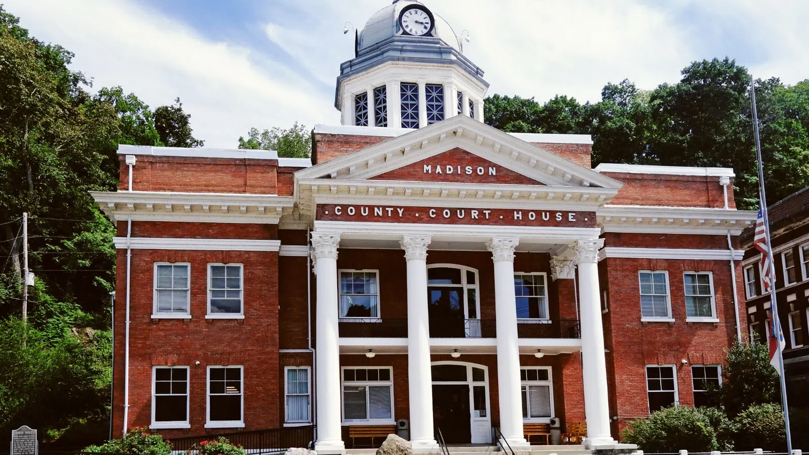 Historic red brick Madison County Courthouse with white columns, clock tower, and lush green trees under blue sky.