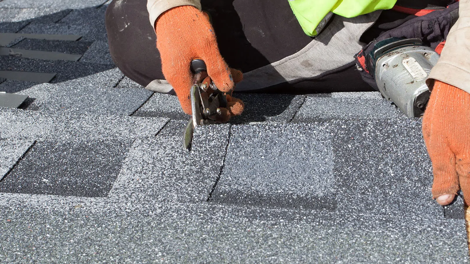 Roofer wearing orange gloves installing asphalt shingles on a roof with cutting tool and nail gun.