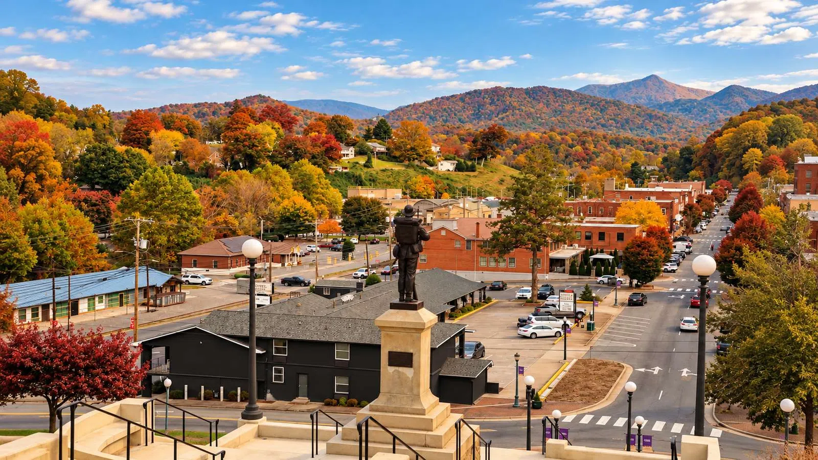 Scenic view of a small town with autumn foliage, statue, historic buildings, and mountains under a blue sky.