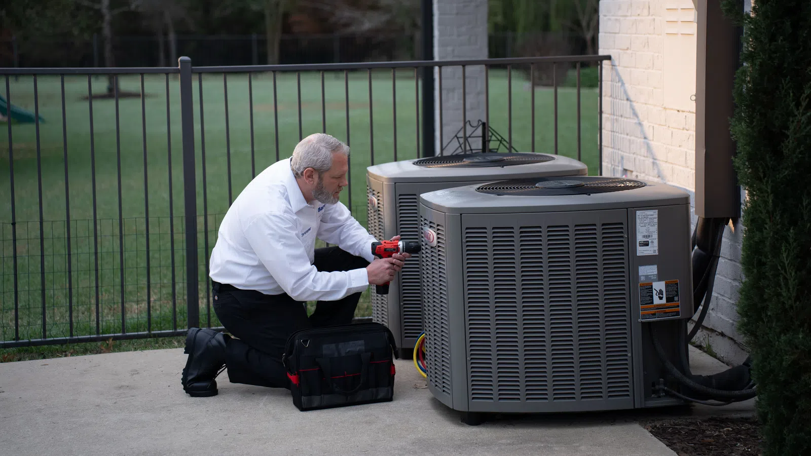 Technician inspecting and repairing two Lennox units beside a house with tools and a toolbox.
