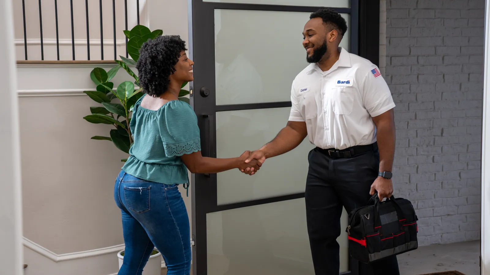 Technician in uniform shaking hands with woman at home entrance, holding tool bag and smiling in a modern hallway.