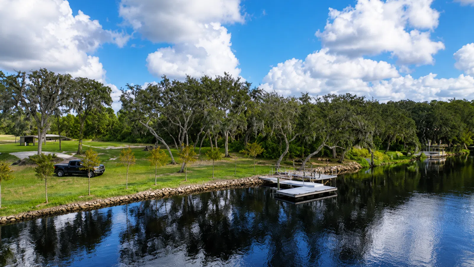 Calm river with tree-lined shore, docks, grassy banks, a parked black truck, and a bright blue sky with clouds.