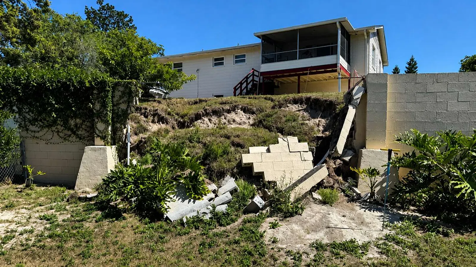 Damaged retaining wall collapsed near grassy backyard slope beneath a house with blue sky above.