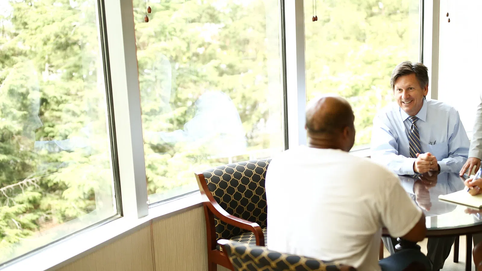 Businessmen having a meeting in a bright conference room with large windows overlooking trees.