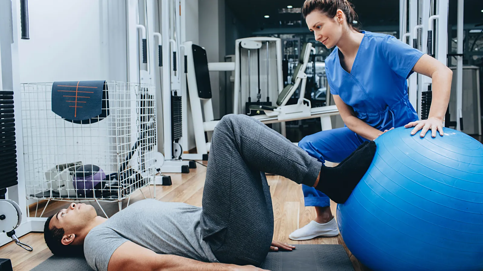 Physical therapist assists man exercising legs using a blue stability ball in a gym setting.