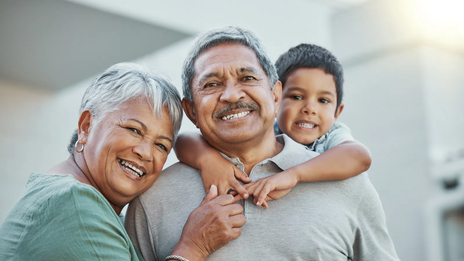 Smiling grandparents with grandson hugging them outdoors in warm sunlight, showing family love and bonding.