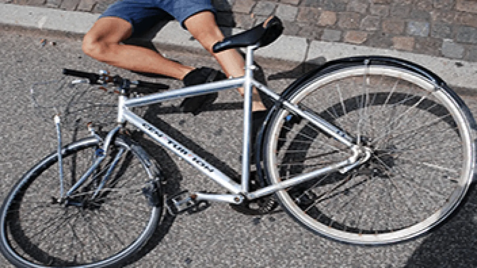Person sitting on pavement next to a silver bicycle lying on its side on a sunny day.