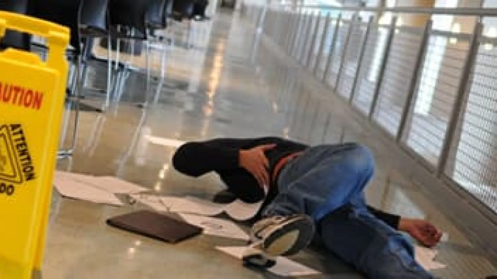 Person lying on floor with scattered papers near caution wet floor sign in indoor hallway area.