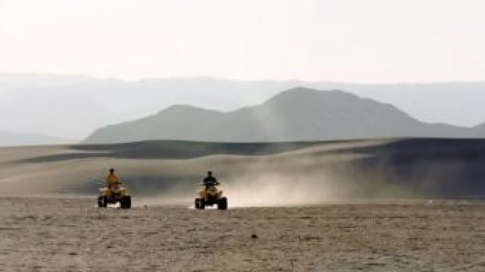 Two people riding ATVs on dusty desert terrain with mountains in the background under a clear sky